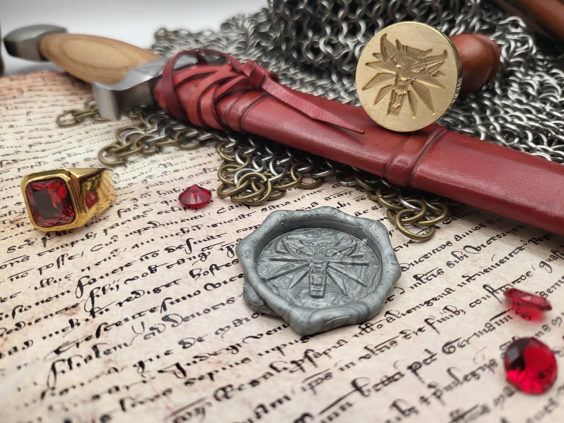A styled flat-lay of fantasy-themed accessories on aged parchment paper, a silver wax seal, a gold ring with a red gem, and chainmail.
