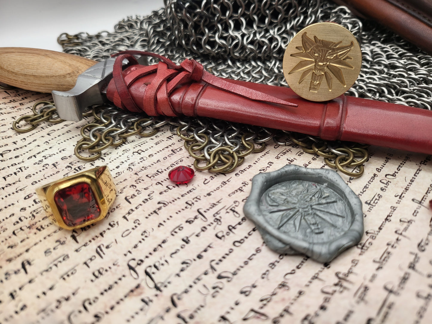 A styled flat-lay featuring a red leather scabbard with a wooden handle, a gold-toned ring with a red gemstone, a silver wax seal, and a small red bead, arranged on aged script paper with chainmail and a hammer.
