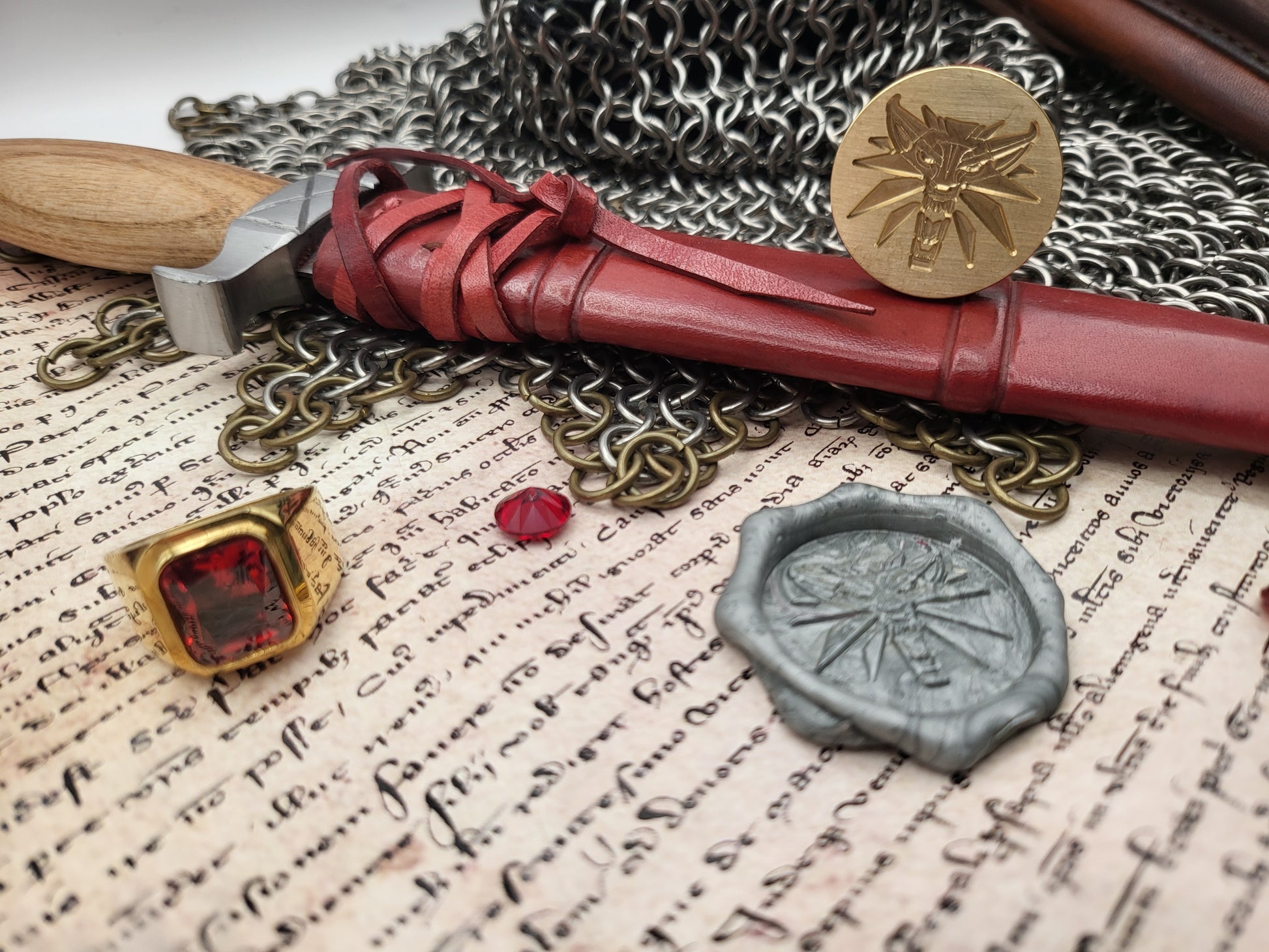 A styled flat-lay featuring a red leather scabbard with a wooden handle, a gold-toned ring with a red gemstone, a silver wax seal, and a small red bead, arranged on aged script paper with chainmail and a hammer.