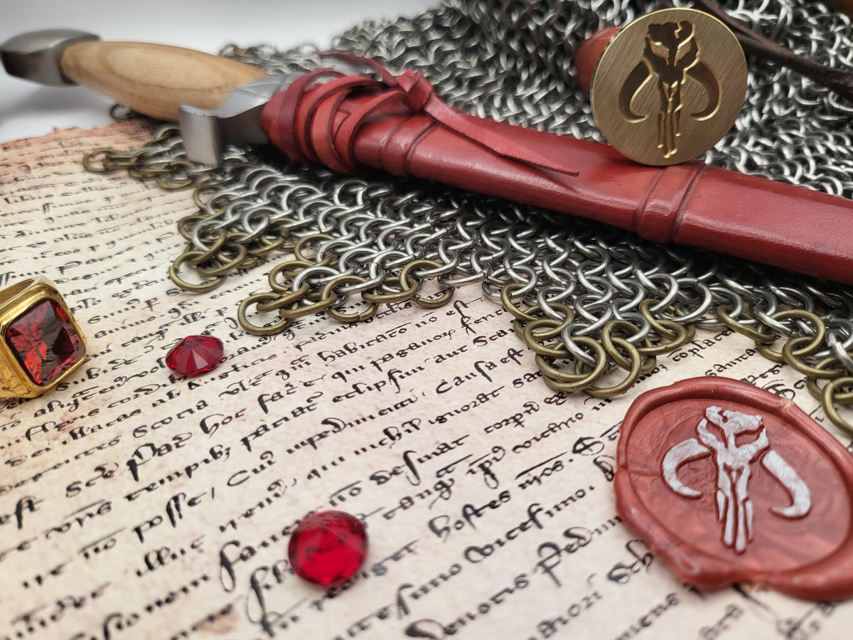 A styled flat-lay of medieval-themed accessories on aged parchment paper, including a red leather-wrapped with a wooden handle, a silver chainmail necklace, a red wax seal stamp, a red wax seal, a gold ring, and a red gemstone.