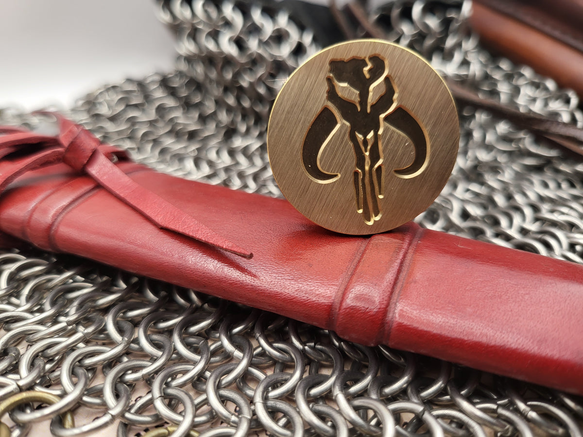 A round brass pendant or medallion with an engraved emblem, displayed on top of a red leather scroll sleeve over a silver chainmail background.