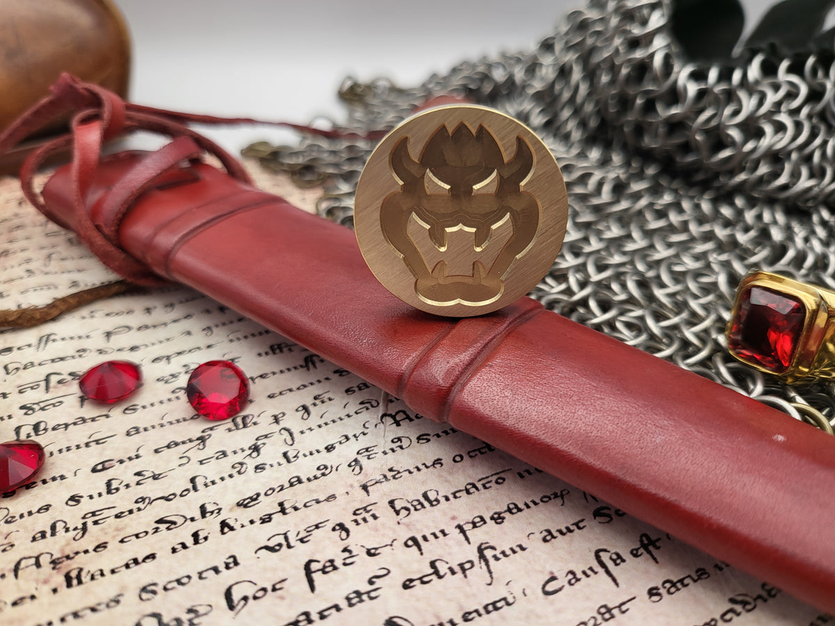 A close-up of a round gold seal stamp with a dark demon face, resting on a red leather scroll sleeve over an old handwritten manuscript, with red gemstones and silver chainmail in the background.