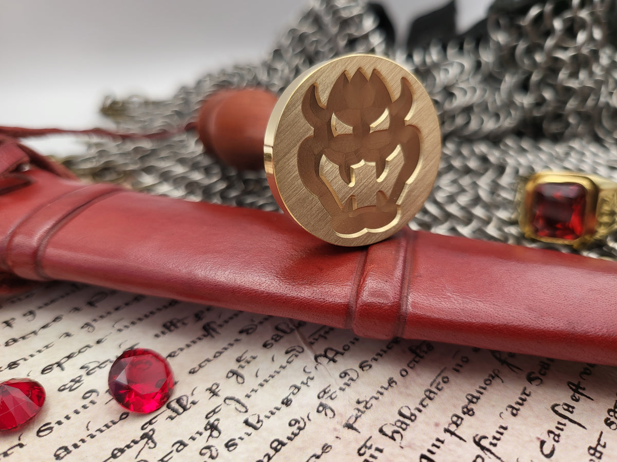 A round brass seal stamp with a carved demon face, resting on a red leather scroll case over an old handwritten manuscript, with red gemstones and a chainmail ring in the background.