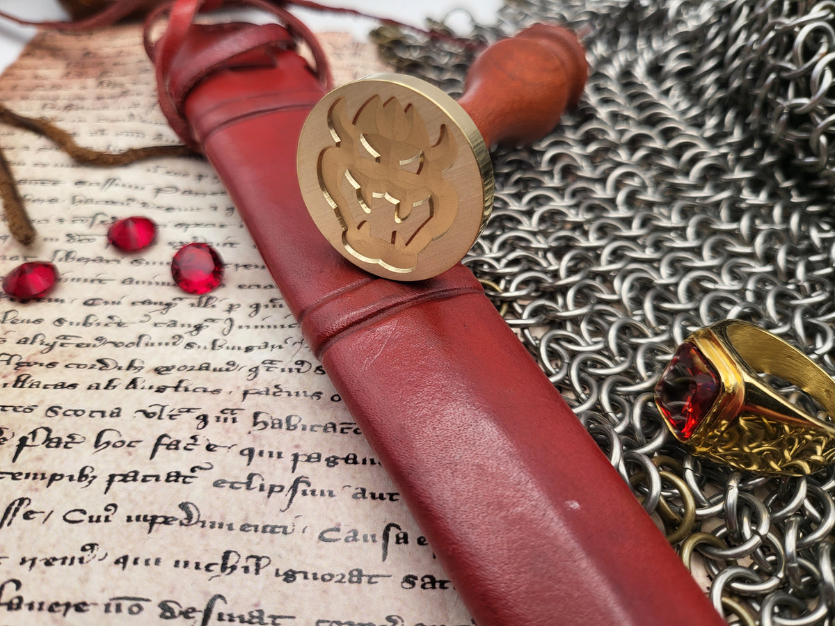 A close-up of a red leather-wrapped seal stamp with a brass seal head, arranged on parchment paper with chainmail and a gold ring with a red gemstone.