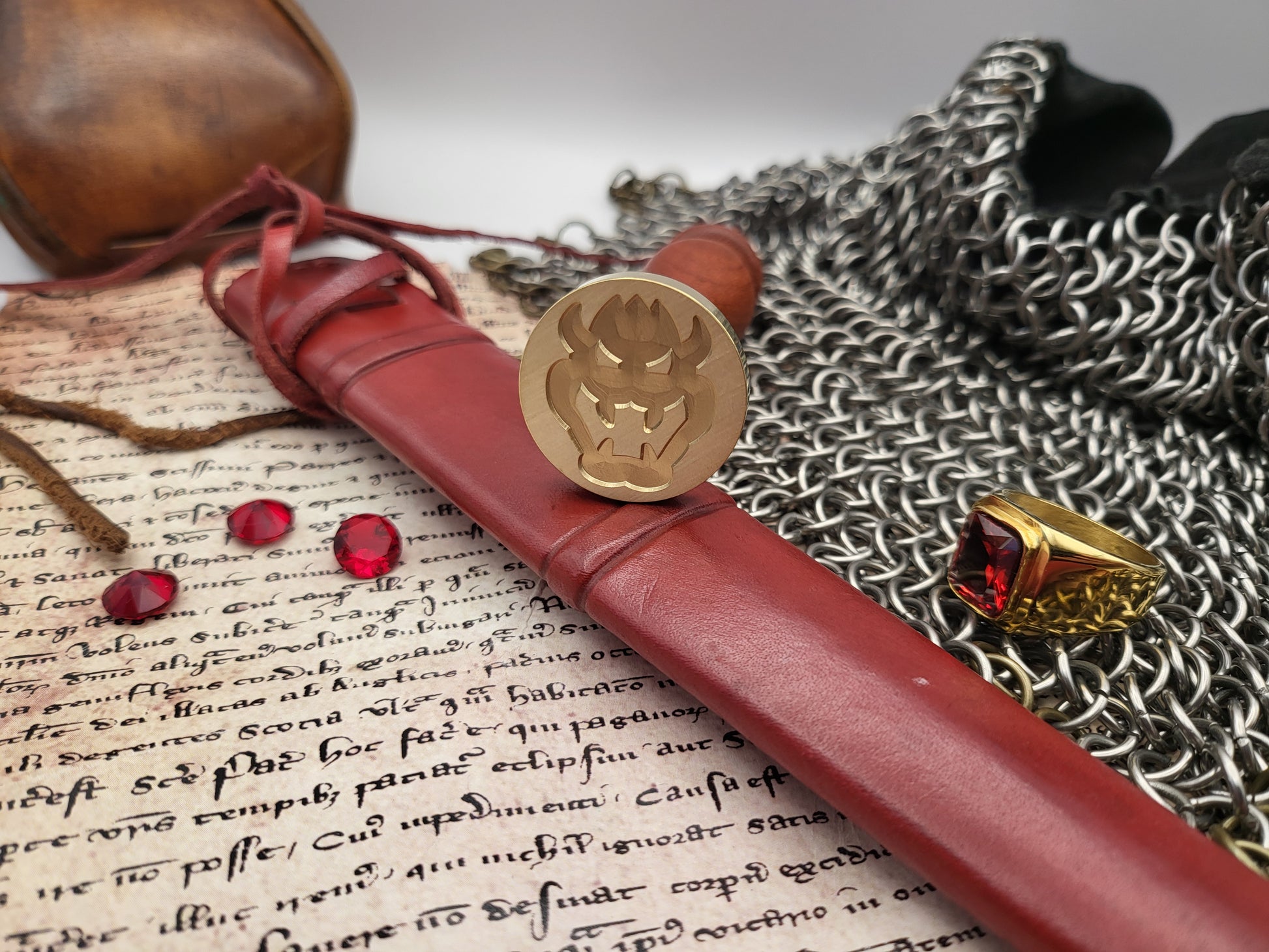 A leather-bound seal with a brass seal stamp resting on an open book, surrounded by medieval-themed props including a ring with a red gem, chainmail, and a leather pouch.