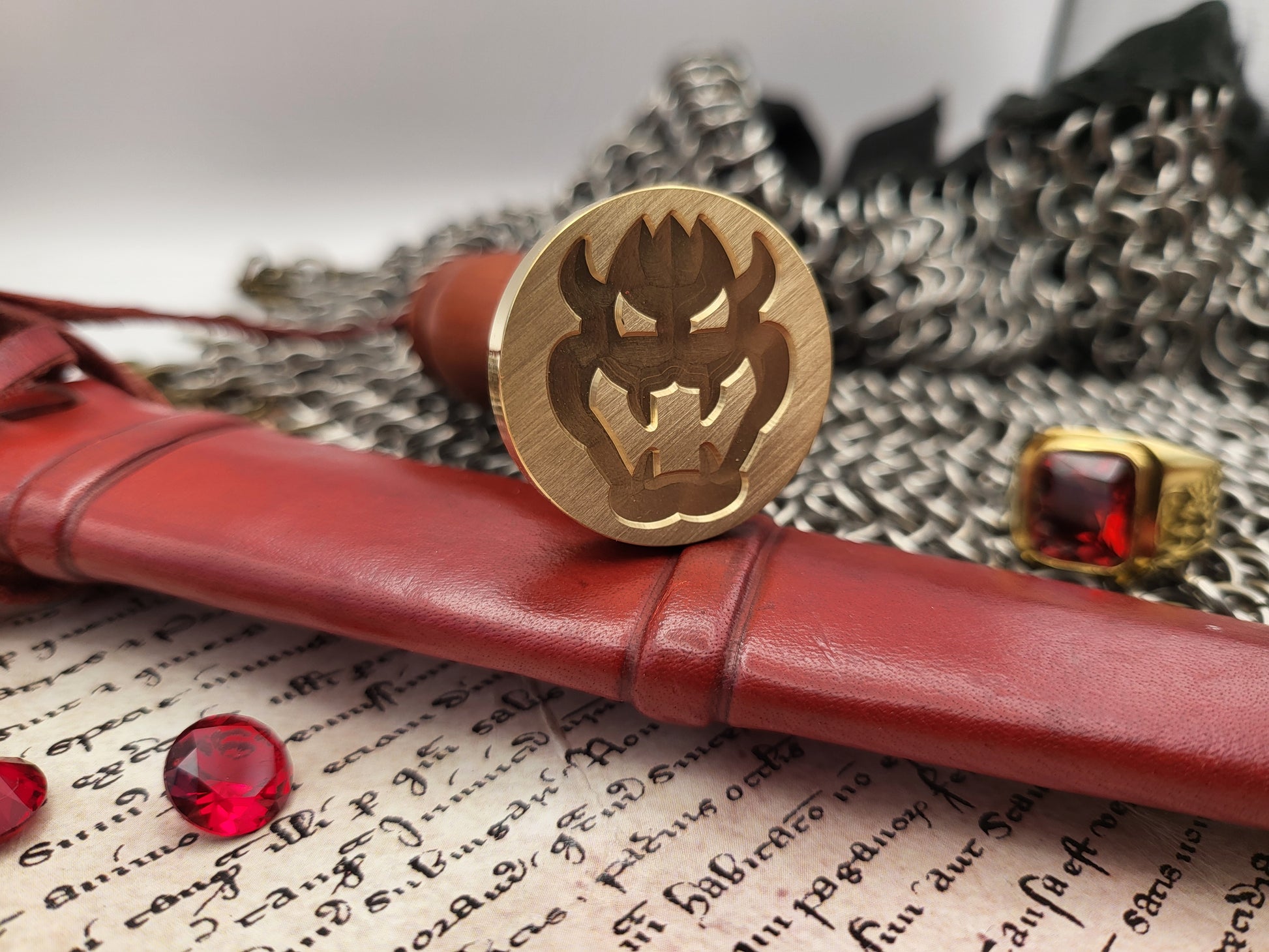 A round gold-toned metal seal or medallion with an engraved horned demon face, displayed on a red leather scroll case over an open book, with chainmail and a red gemstone ring in the background.