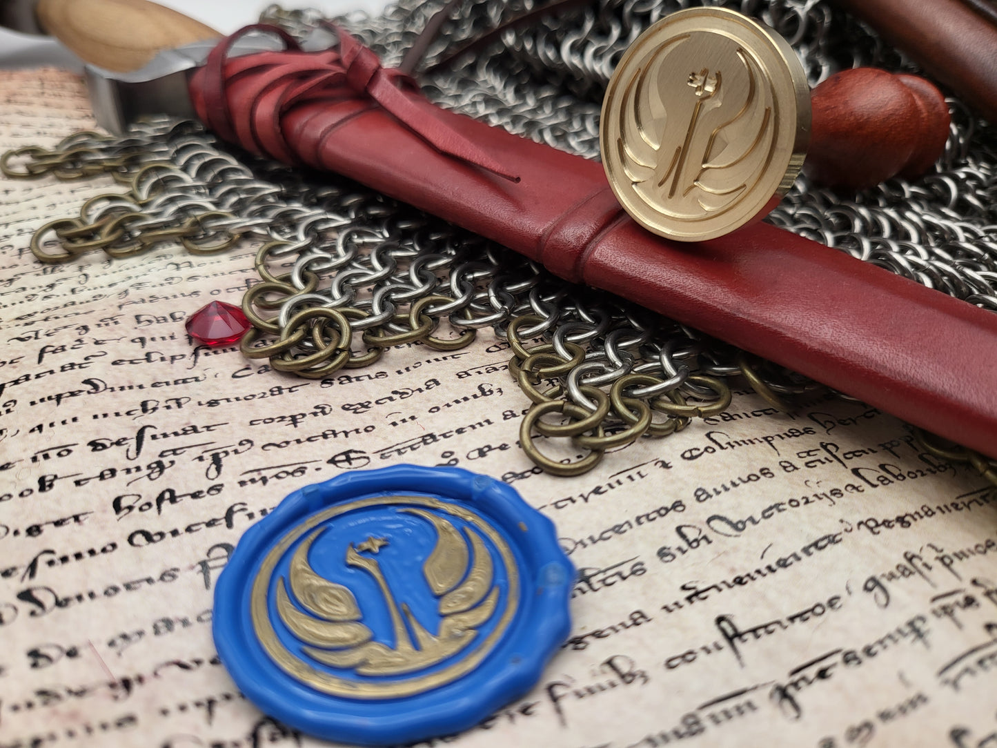 A close-up styled product shot showing a red leather seal wax envelope tied with a cord, a round gold wax seal, a blue wax seal with a gold emblem, chainmail, and aged script paper.