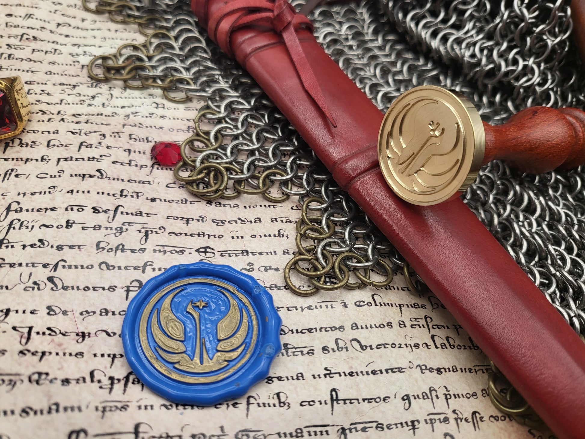 A close-up styled product shot showing a red leather seal wax envelope, a red wax seal stamp with a gold emblem, a blue wax seal stamp with a gold emblem, and a chainmail accessory laid over an aged handwritten manuscript.
