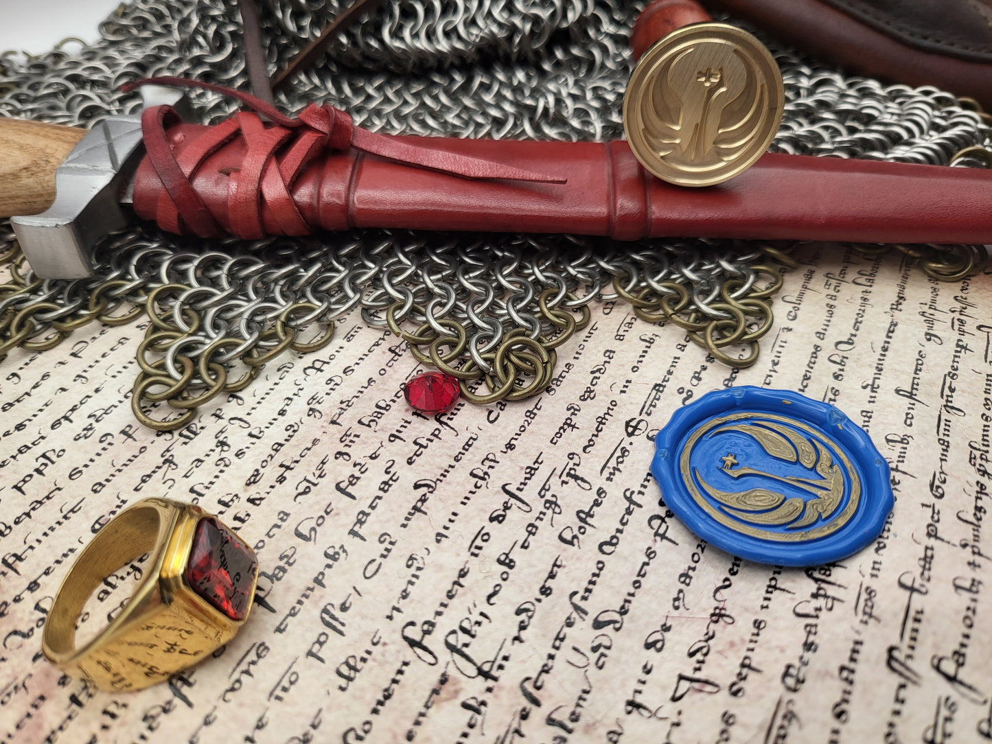 A styled flat-lay of medieval-themed accessories on aged parchment paper, a blue wax seal, a gold ring with a red stone, a brass token, and a small red gem on a chainmail backdrop.