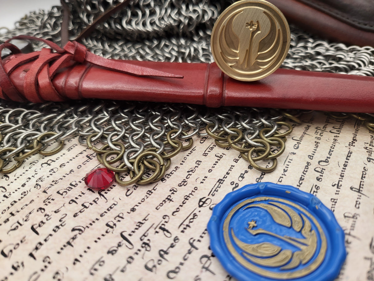 A close-up styled arrangement featuring a red leather-wrapped scroll with a gold seal, a blue wax seal with a gold emblem, chainmail, and aged handwritten paper.