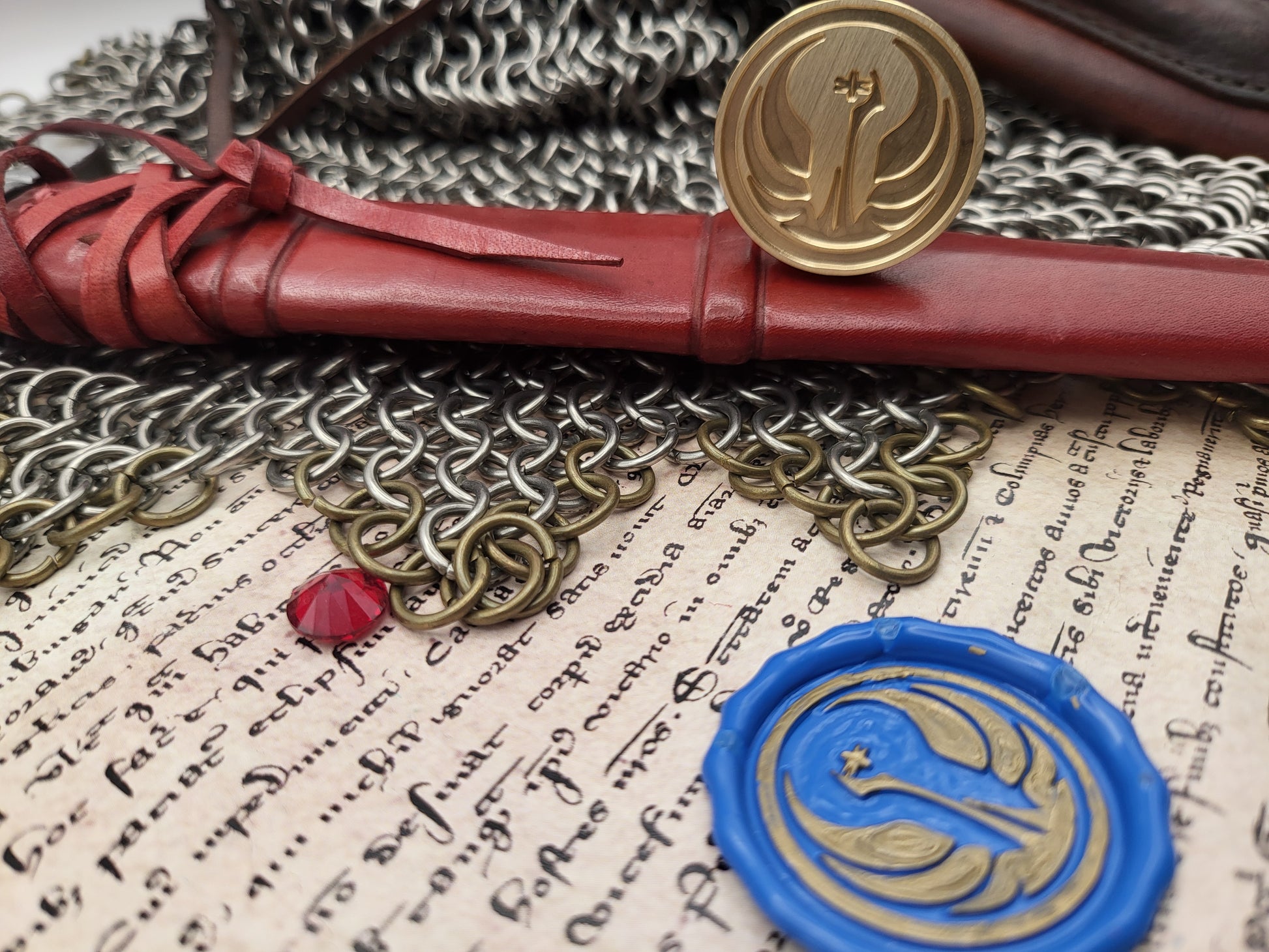 A close-up styled arrangement featuring a red leather-wrapped scroll with a gold seal, a blue wax seal with a gold emblem, chainmail, and aged handwritten paper.