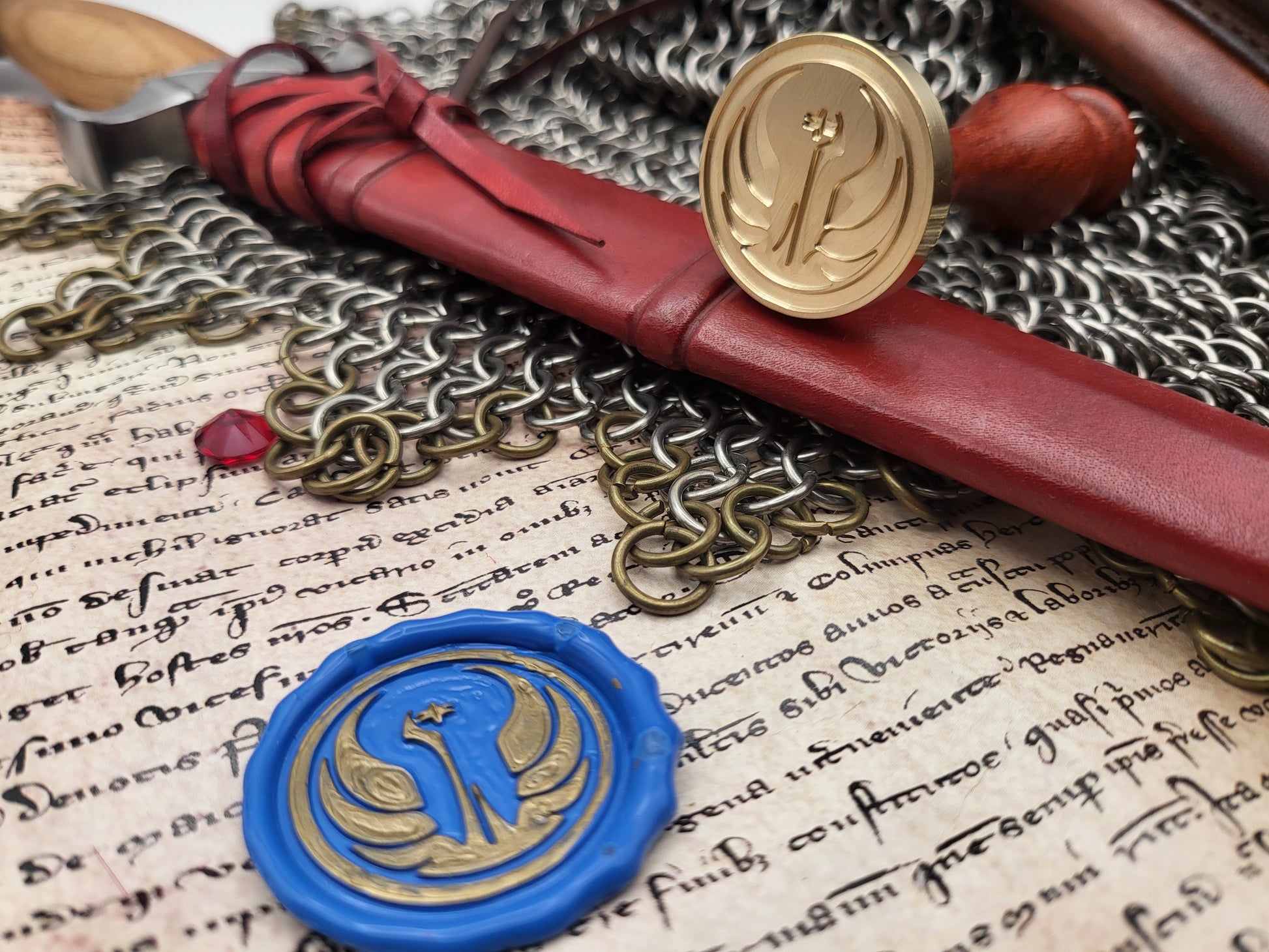 A close-up styled arrangement featuring a red leather seal stamp with a gold emblem, a blue wax seal stamp with a gold emblem, chainmail, parchment with cursive writing, and a small wooden object.