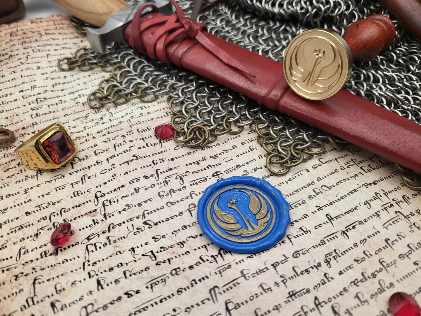 A styled flat-lay of medieval-themed accessories on aged parchment paper with handwritten script, including a red leather scroll seal with a gold emblem, a blue wax seal with a gold emblem, chainmail, a ring, and a sword hilt.