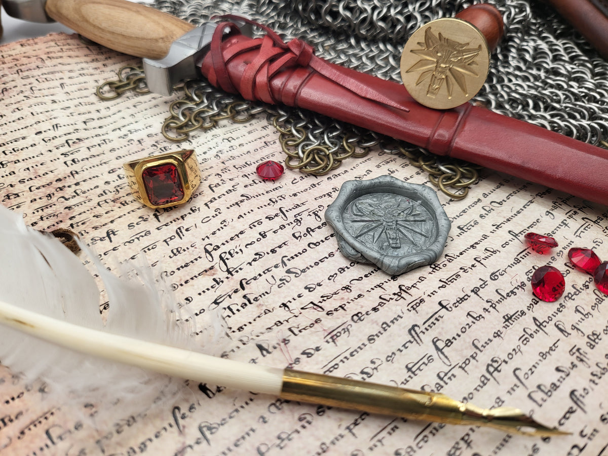 A styled flat-lay of medieval-themed accessories on aged script paper, including a red wax seal stamp with a gold bee emblem, a silver wax seal, a gold ring with a red gem, a red wax seal, a feather quill with a gold-tipped pen, chainmail, and a wooden-handled tool.